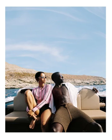 A couple sits on a boat with a rocky shoreline visible in the background. The woman wears sunglasses and a pink shirt, the man wears a white shirt.