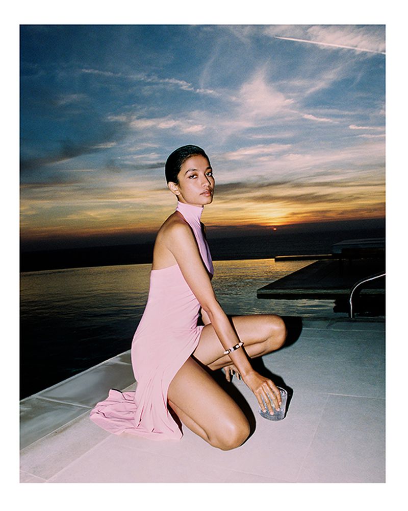 Woman in pink dress poses poolside at sunset. She kneels holding a drink with an ocean and sky backdrop. Fashion shoot, dusk lighting.