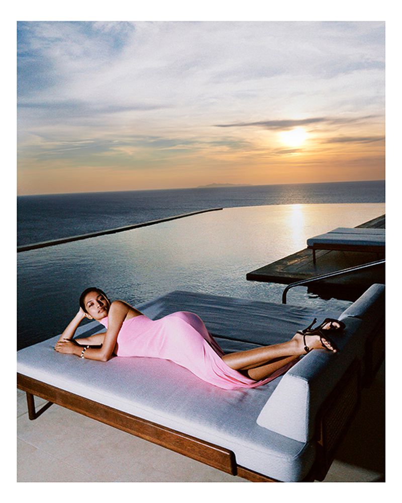 A woman in a pink dress is reclining on a lounge chair near an infinity pool overlooking a sunset view of the ocean.