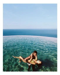 Man holding a woman in an infinity pool overlooking the ocean under a clear sky.