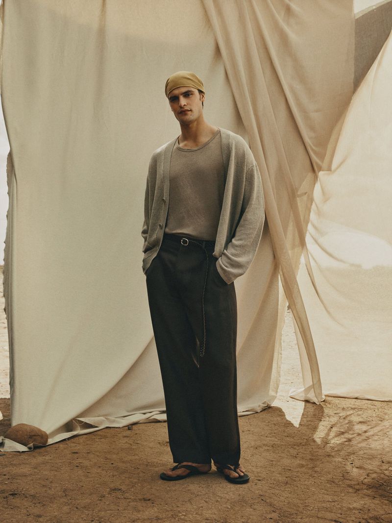 A full shot of a male fashion model posing in a tan shirt, cardigan, and black pants in front of a draped backdrop.