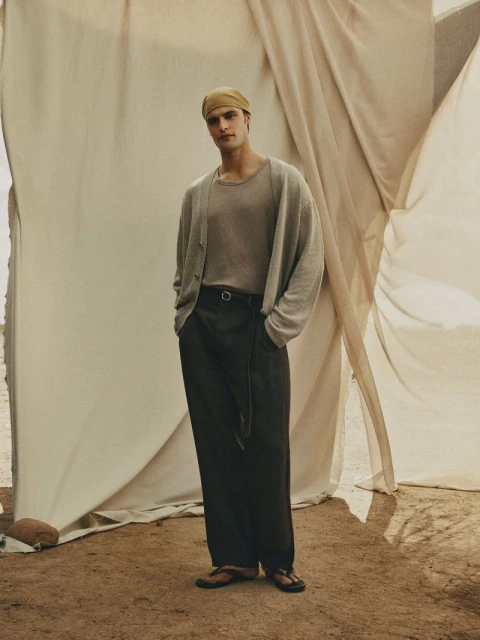 A full shot of a male fashion model posing in a tan shirt, cardigan, and black pants in front of a draped backdrop.