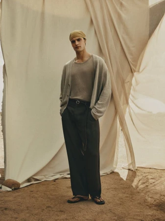 A full shot of a male fashion model posing in a tan shirt, cardigan, and black pants in front of a draped backdrop.