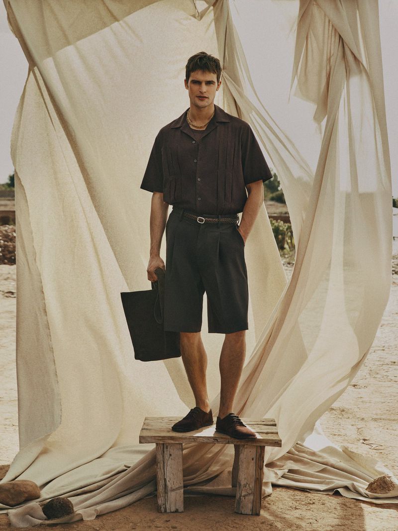 A man stands on a wooden stool holding a black tote bag, wearing a dark brown short-sleeved shirt, dark shorts, and dark shoes.