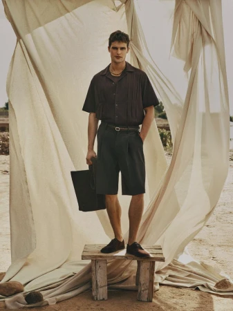 A man stands on a wooden stool holding a black tote bag, wearing a dark brown short-sleeved shirt, dark shorts, and dark shoes.