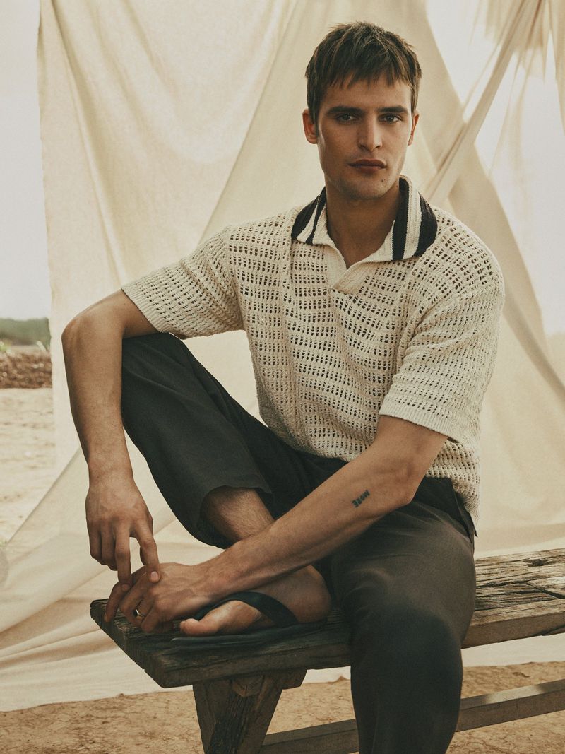 A relaxed adult male sits on a wooden bench wearing a knitted polo shirt and dark pants, against a backdrop of draped fabric.