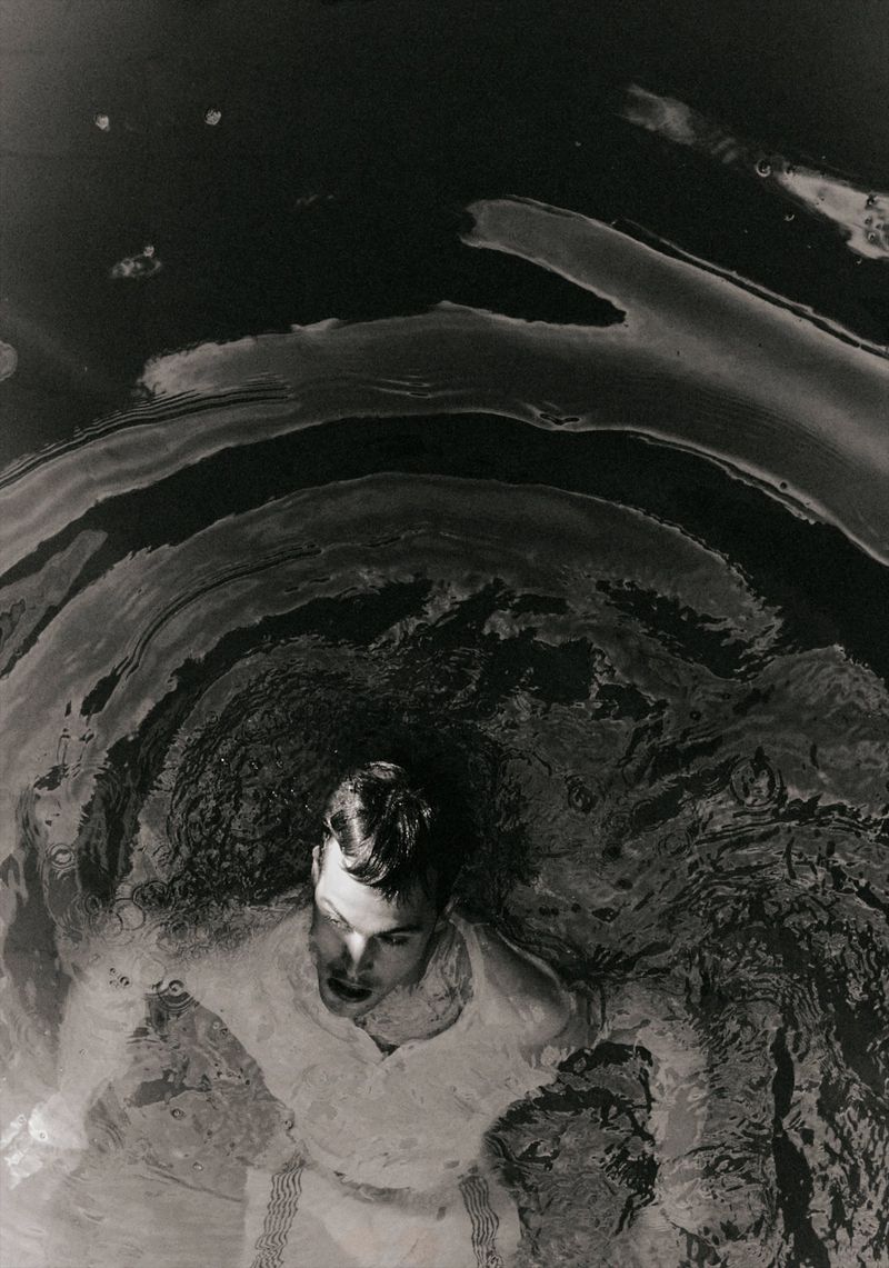 Monochrome portrait of an adult male partially submerged in water, looking up with his mouth open in an outdoor environment.