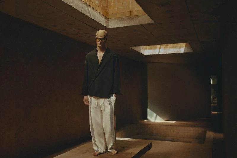 An adult male stands on a platform in a dark, indoor space wearing a black shirt, light pants, turban, and eyeglasses, with a solemn expression.