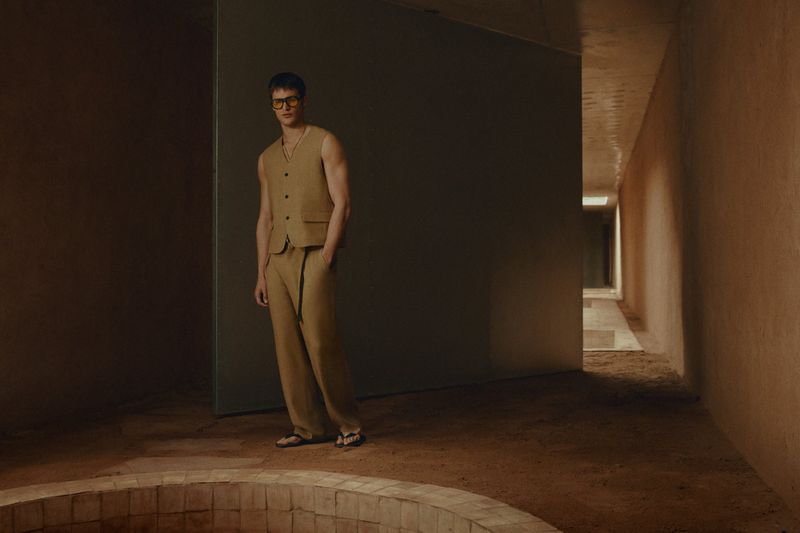 A slender man poses indoors, wearing a vest and sunglasses, with his hand casually placed in his pocket, set against a modern, earthy backdrop.