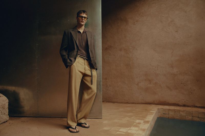 Man posing in brown jacket and tan pants, standing in front of a metal wall, indoor setting with a minimalist aesthetic.