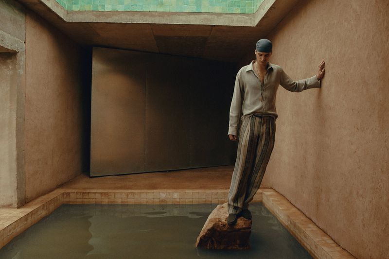 Young adult male poses in relaxed style inside a pool-surrounded room with light gray shirt and striped pants in soft lighting.