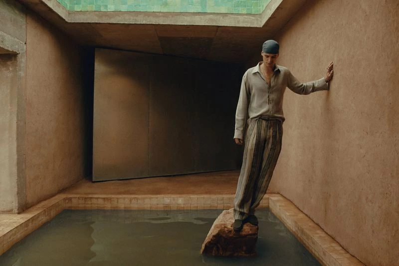 Young adult male poses in relaxed style inside a pool-surrounded room with light gray shirt and striped pants in soft lighting.
