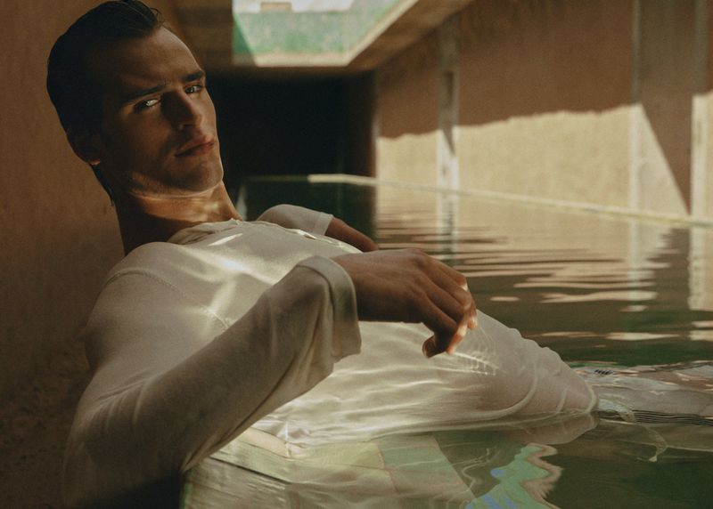 Adult male model with fair skin wearing a white long-sleeve shirt lounging in a pool surrounded by a tan-colored building structure.