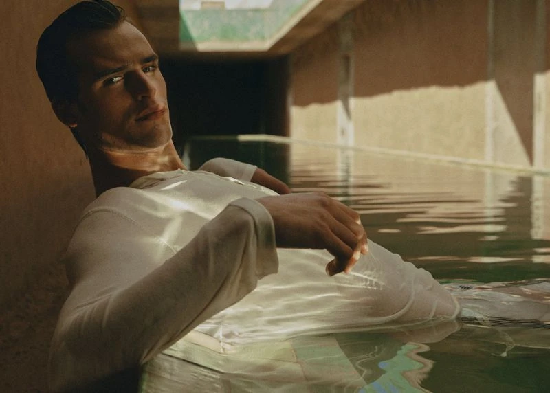 Adult male model with fair skin wearing a white long-sleeve shirt lounging in a pool surrounded by a tan-colored building structure.