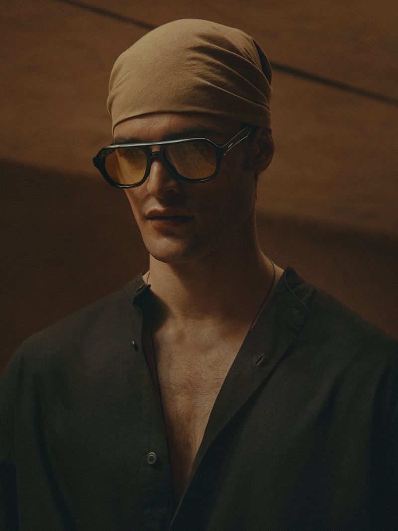 Portrait of a man wearing dark glasses and a tan headscarf. He is wearing a button-up shirt with an open collar in a studio setting.