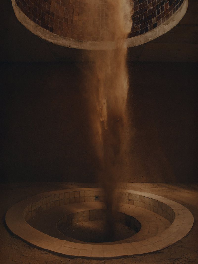Abstract visual featuring a tan material, like sand, cascading from a circular opening onto a spiral structure in a dark room.