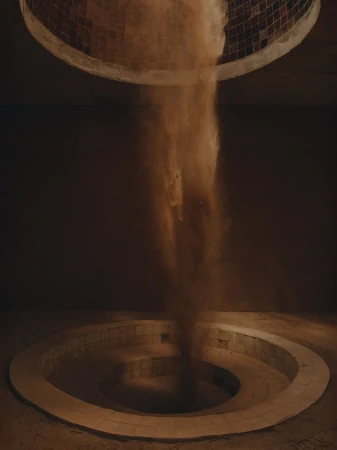 Abstract visual featuring a tan material, like sand, cascading from a circular opening onto a spiral structure in a dark room.