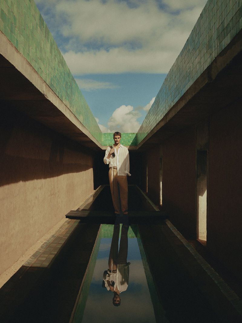 A stylish man in a white shirt and tan pants stands confidently on a narrow platform above reflecting water within a high-walled corridor.