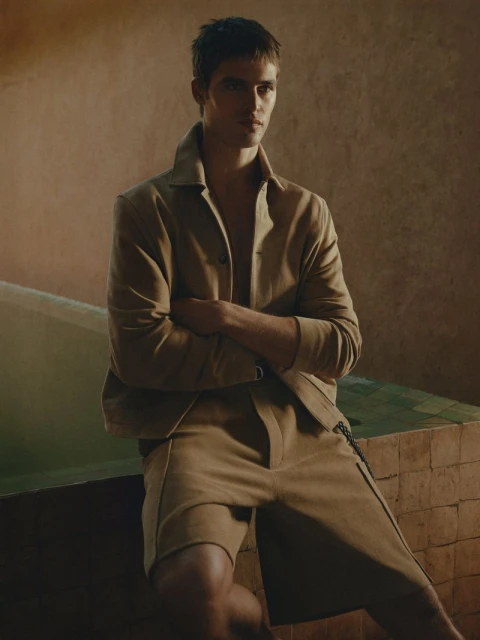 A fair-skinned male model with folded arms poses in a tan jacket and shorts, set against a warm-toned background with a tiled surface.
