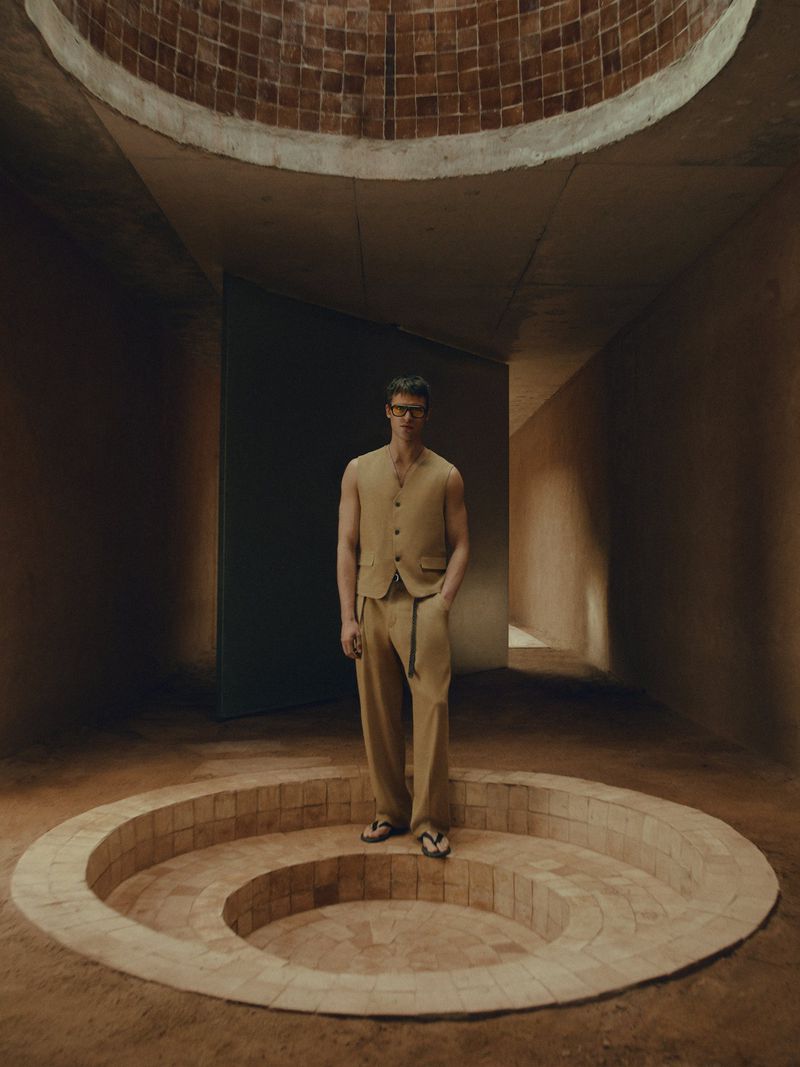 An adult male model in a light brown vest and pants stands on a round terracotta-tiled platform in a minimalist indoor setting.