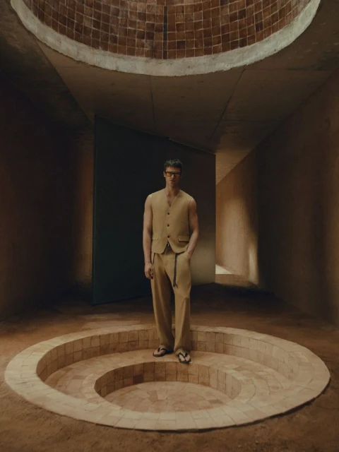 An adult male model in a light brown vest and pants stands on a round terracotta-tiled platform in a minimalist indoor setting.
