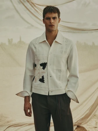 A stylish adult male poses outdoors wearing a white collared jacket with floral embroidery and dark trousers, set against a draped backdrop.