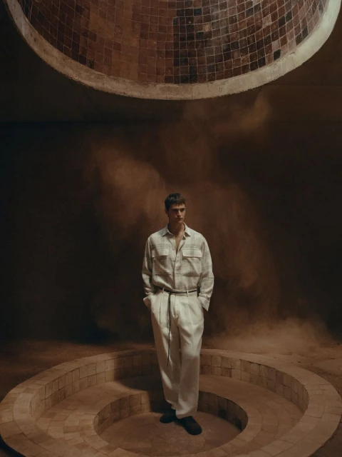 A casually dressed man in an earthy-toned indoor fashion shot, featuring diffused lighting, architectural background details, and a contemplative mood.