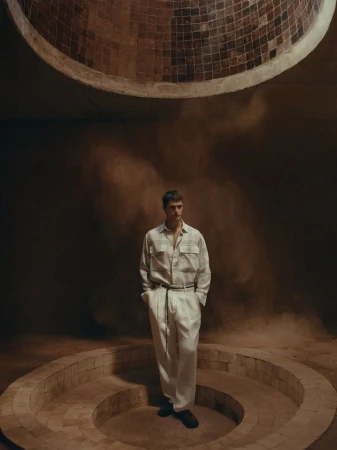 A casually dressed man in an earthy-toned indoor fashion shot, featuring diffused lighting, architectural background details, and a contemplative mood.