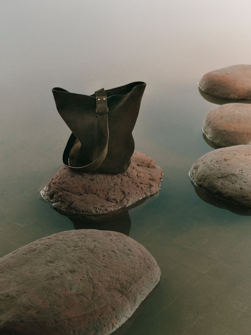 Leather tote bag is displayed on a rock in standing water, creating a minimalist and natural scene with a calm and muted color palette.