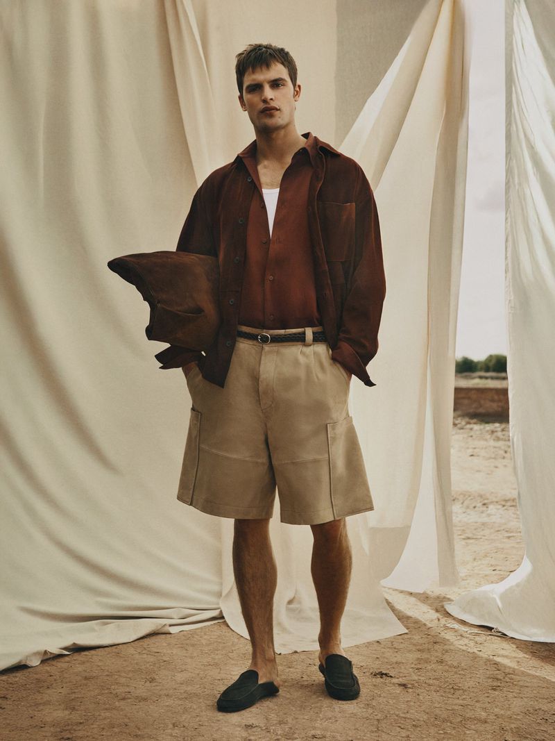 Full-length portrait of a handsome male fashion model outdoors wearing a brown shirt, khaki shorts, and black loafers.