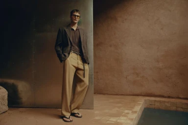 Male model wearing a brown blazer, polo shirt, and beige trousers standing by a metallic panel next to a pool.