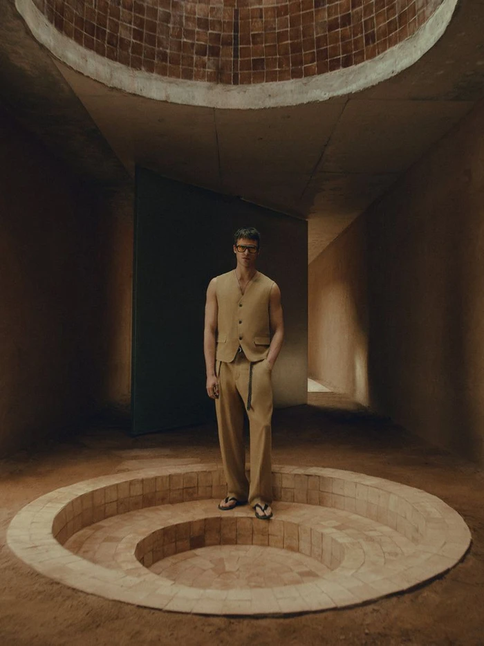 Parker Van Noord in a beige vest and trousers standing inside a rustic architectural interior with a circular tiled sunken pit.