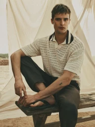 Parker Van Noord in a cream knit polo shirt and dark trousers sitting on a wooden bench against fabric backdrop for Zara.