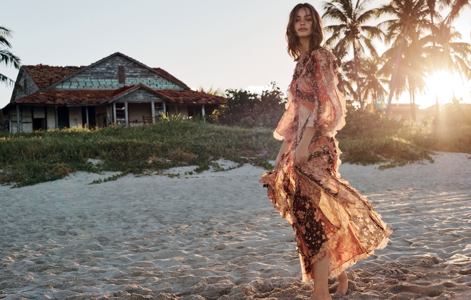 Woman in a flowing dress posing on a sandy beach with palm trees and an old weathered house in the background at sunset.