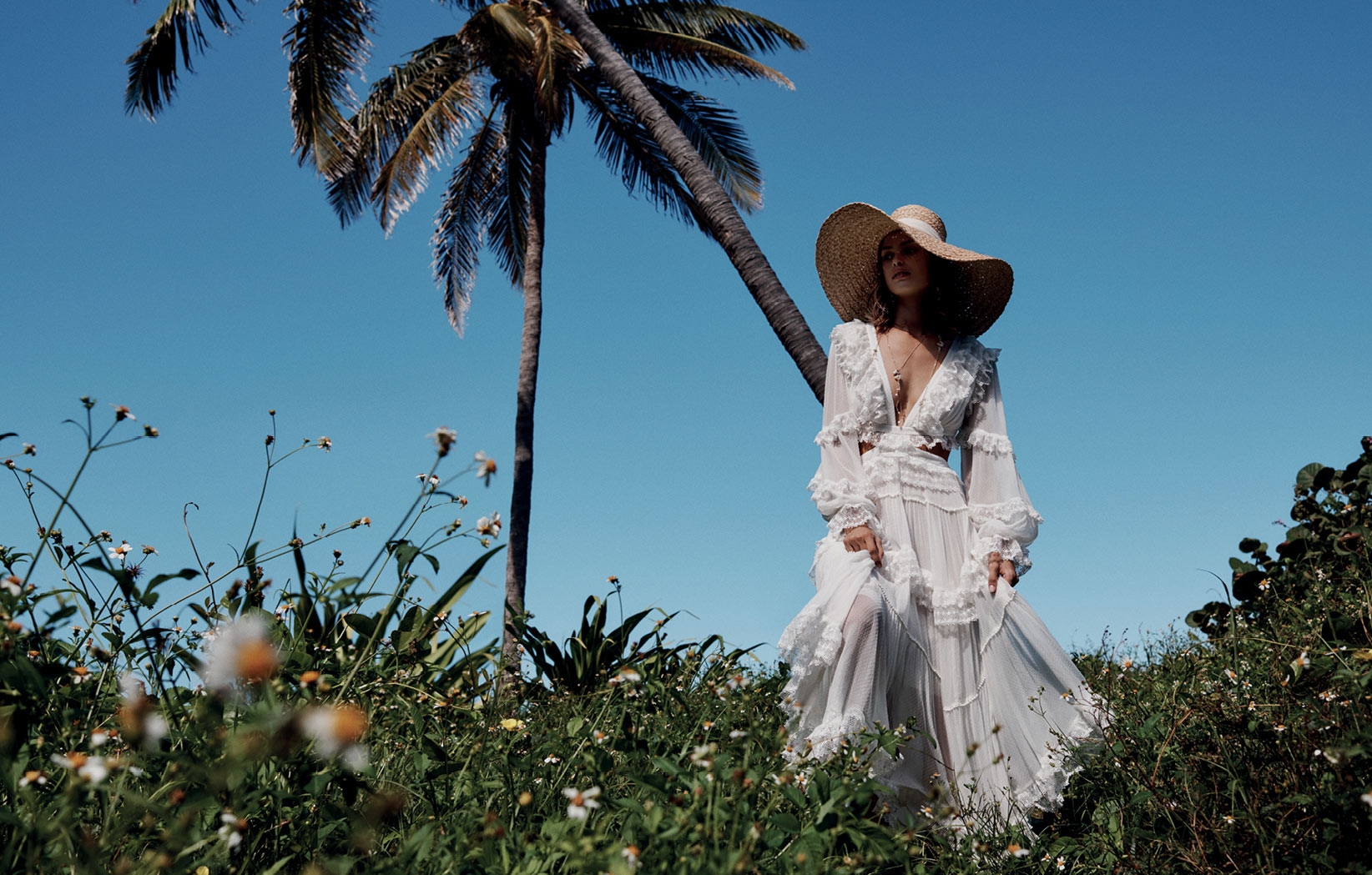 A woman wearing a white dress and straw hat poses outdoors near a palm tree under a blue sky, surrounded by green plants.