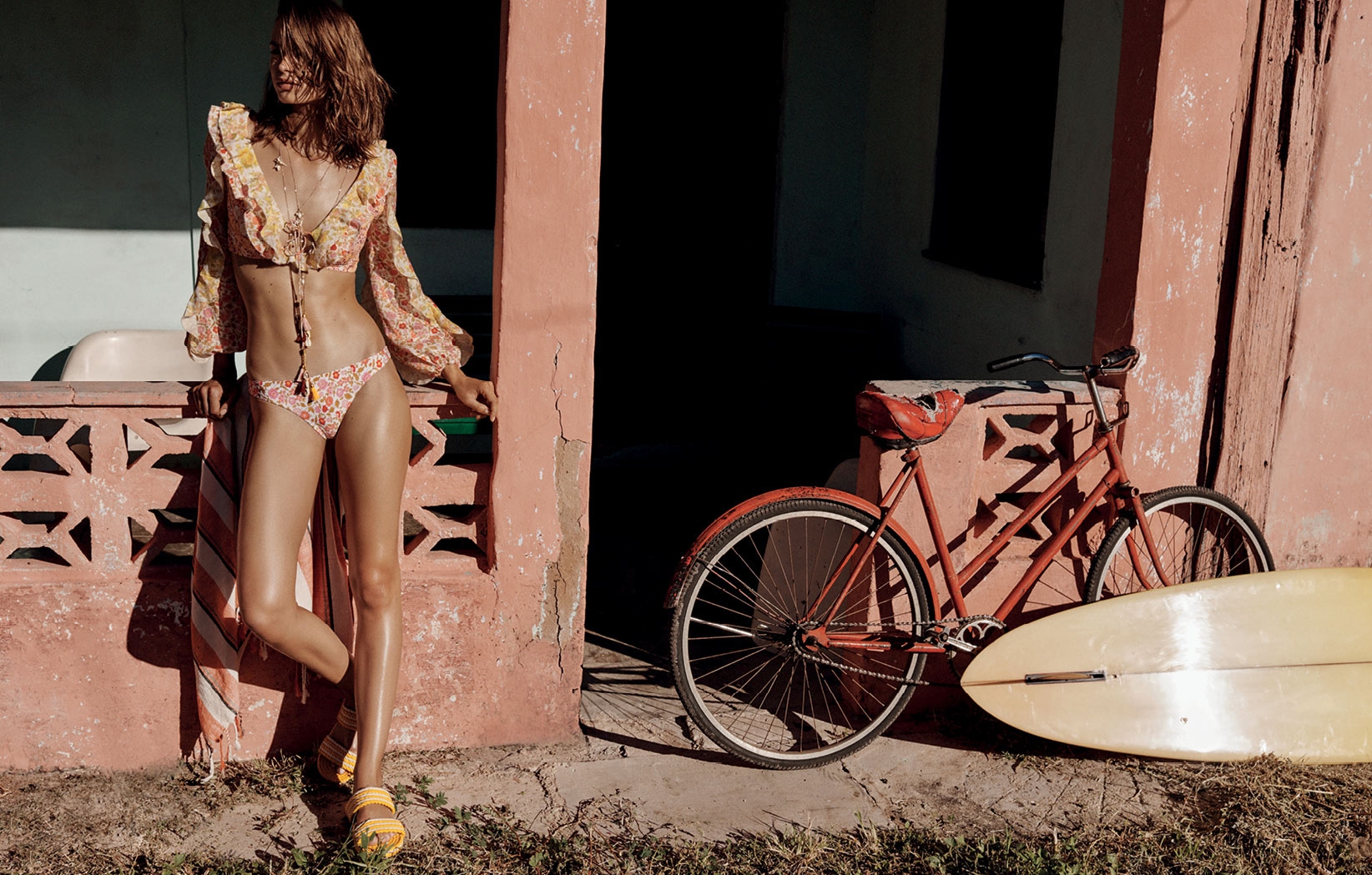 Adult woman poses in floral bikini with a vintage bicycle and surfboard against a coral wall, warm sunny light, summer aesthetic.