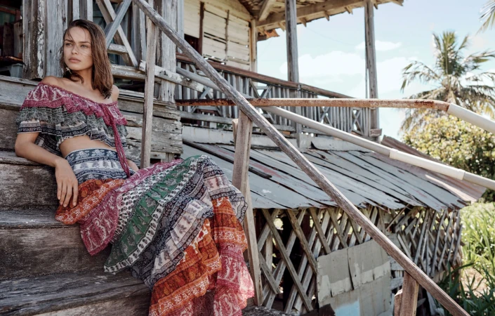 Adult woman wearing patterned crop top and skirt sitting on wooden steps outside of dilapidated building under partly cloudy sky.
