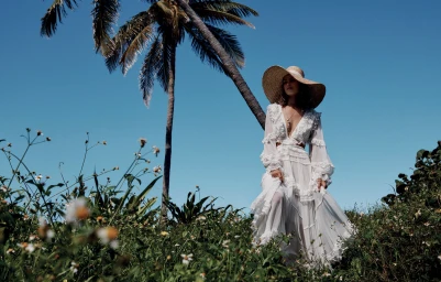 Model in a long white ruffled dress and wide-brimmed straw hat standing among green foliage with a palm tree and clear blue sky.