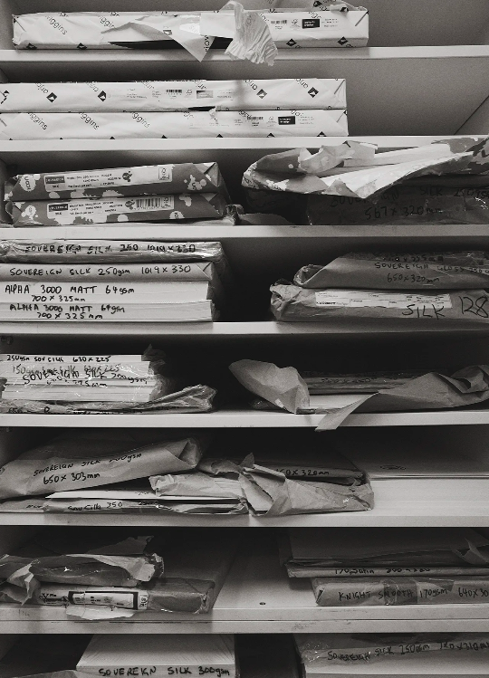 Shelves hold reams of paper wrapped in paper, some with handwritten labels in a grayscale photo; an organizational system, printing supplies.