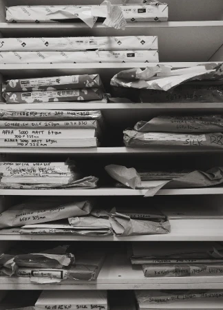 Shelves hold reams of paper wrapped in paper, some with handwritten labels in a grayscale photo; an organizational system, printing supplies.