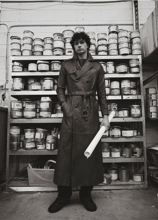 Monochrome portrait of a man in a trench coat standing in front of shelves stocked with cans, holding a white rolled-up tube.
