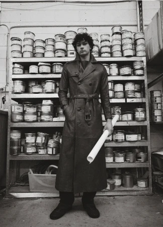 Monochrome portrait of a man in a trench coat standing in front of shelves stocked with cans, holding a white rolled-up tube.