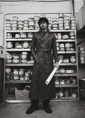 Man wearing a long dark belted coat standing in front of metal shelving stacked with circular containers