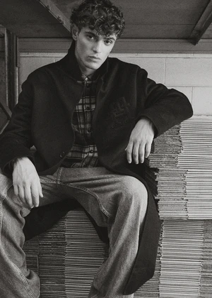 A young male model with curly hair seated and leaning on stacked cardboard boxes wearing a dark coat and plaid shirt.
