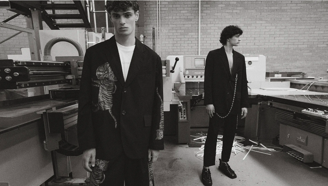 Two male fashion models wear graphic print suits in a monochrome industrial setting, one with a tiger design. Editorial style.
