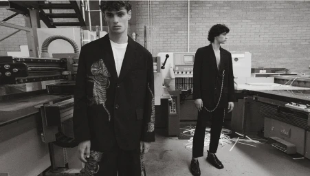 Two male models in dark suits standing inside a factory workshop with machinery and brick walls