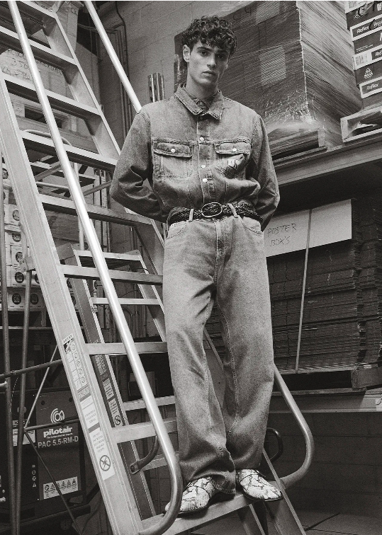 Monochromatic fashion shot of a young adult male posing on metal steps in a denim outfit with boxes in the background.