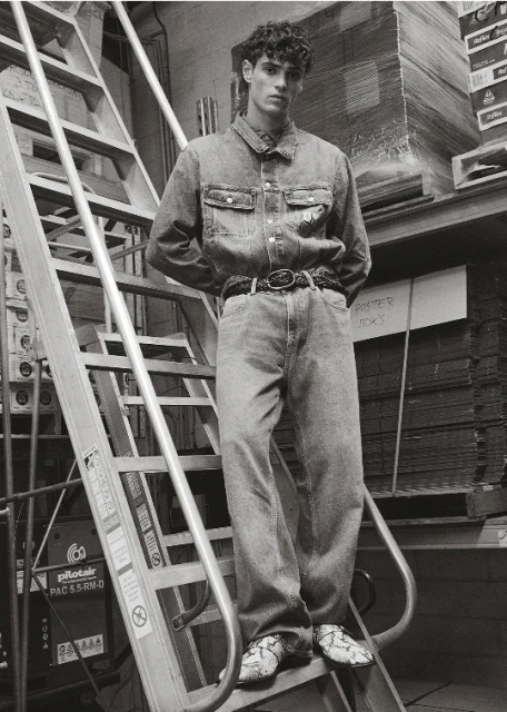 Monochromatic fashion shot of a young adult male posing on metal steps in a denim outfit with boxes in the background.