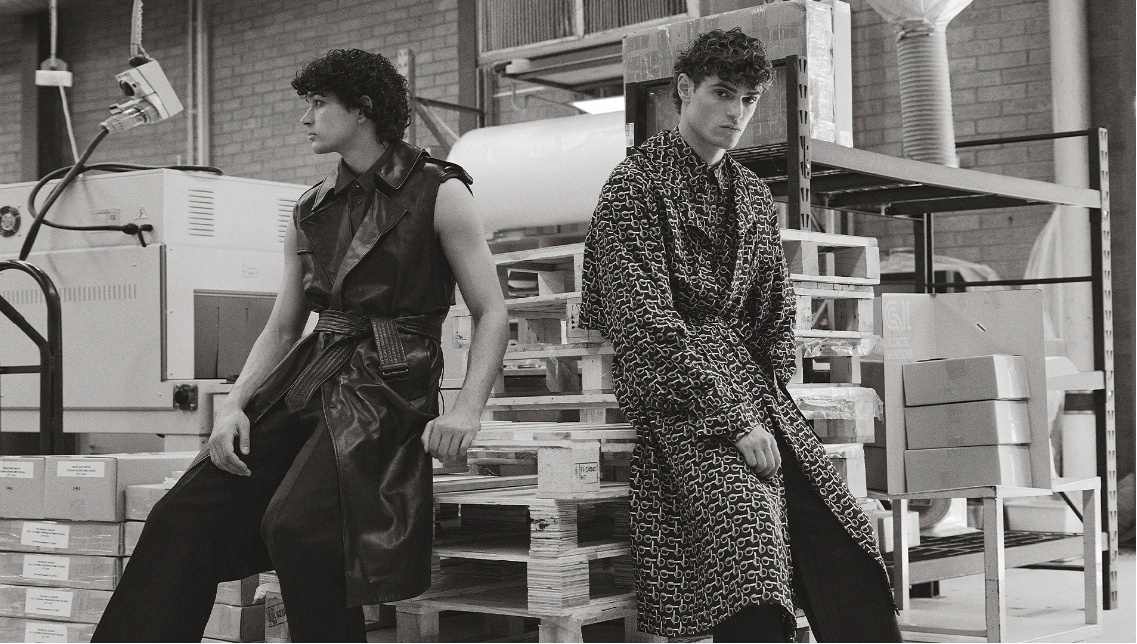 Two adult men in patterned coats, posing on a wooden pallet in an industrial setting, creating a high-fashion editorial image.