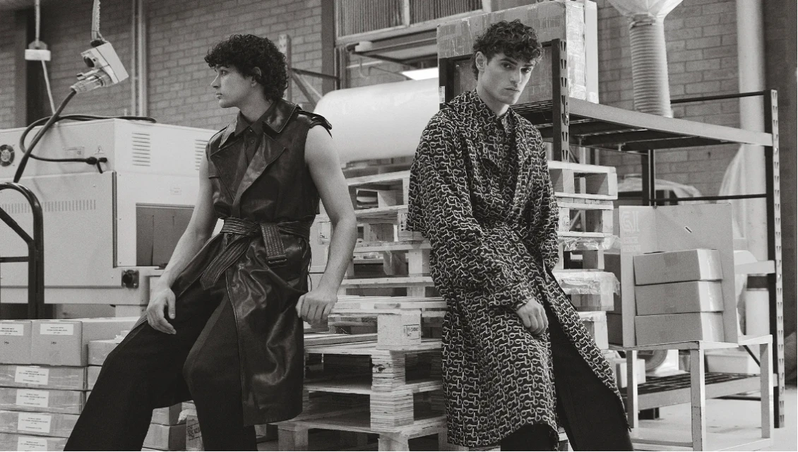 Two adult men in patterned coats, posing on a wooden pallet in an industrial setting, creating a high-fashion editorial image.